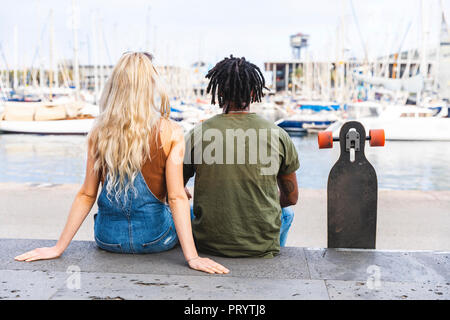 Spanien, Barcelona, Rückansicht des multikulturellen junges Paar sitzen Seite an Seite mit Longboard im Marina Stockfoto