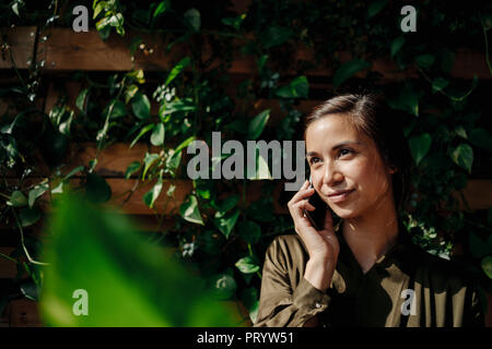 Lächelnde junge Frau auf Handy an der Wand mit Kletterpflanzen Stockfoto