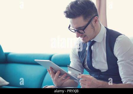 Jungen gutaussehenden Mann in formalen Outfit und Gläsern sitzen auf einem Sofa in modernes Apartment mit Tablet mit Kreditkarte Stockfoto