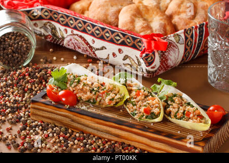Mediterraner Bulgur / Couscous Salat serviert in Salatblättern und Kirschtomaten auf einem Holzbrett. Eine gesunde vegane Mahlzeit. Stockfoto