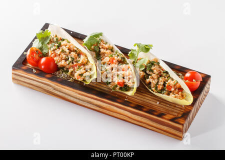 Mediterraner Bulgur / Couscous Salat serviert in Salatblättern und Kirschtomaten auf einem Holzbrett. Eine gesunde vegane Mahlzeit. Isoliert auf Weiß. Stockfoto