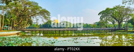 YANGON, MYANMAR - Februar 27, 2018: Panorama der Kandawgyi Park mit alten hölzernen Brücke über den See, bedeckt mit Lotus Pflanzen und umgeben durch sp Stockfoto