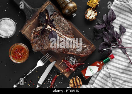 Gegrilltes große T-Bone Steak garniert mit Kräutern, Getreide und Salz mit Messer und Gabel daneben auf Schneidebrett auf schwarzem Hintergrund. Ansicht von oben. Stockfoto