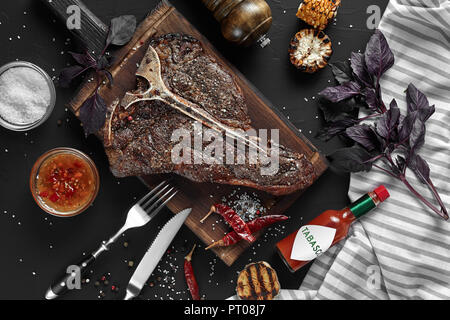 Gegrilltes große T-Bone Steak garniert mit Kräutern, Getreide und Salz mit Messer und Gabel daneben auf Schneidebrett auf schwarzem Hintergrund. Ansicht von oben. Stockfoto