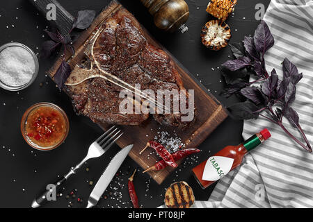 Gegrilltes große T-Bone Steak garniert mit Kräutern, Getreide und Salz mit Messer und Gabel daneben auf Schneidebrett auf schwarzem Hintergrund. Ansicht von oben. Stockfoto