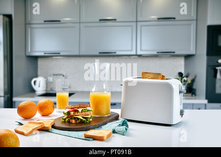 Weiß elektrische Toaster mit Orangensaft mit Sandwich am Küchentisch. Frühstück in der gemütlichen Küche. Nützliches und leckeres Essen Stockfoto