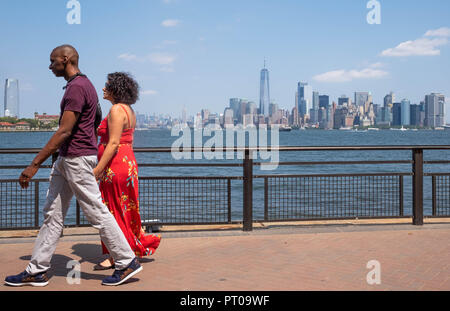 Ein paar gehen Hand in Hand auf Liberty Island mit der New York City Skyline im Hintergrund, einschließlich Lower Manhattan und der Freedom Tower Stockfoto