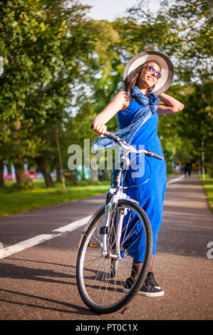 Junge brünette Mädchen im blauen Kleid und hellen Hut Holding ein Fahrrad in einem Park in der Gasse. Stockfoto