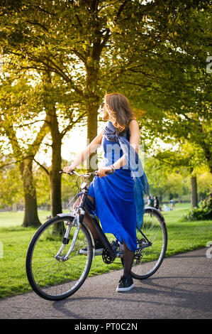 Junge brünette Mädchen im blauen Kleid und Sonnenbrille auf einem Fahrrad Sonnenuntergang Licht.. Stockfoto