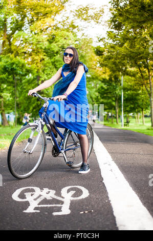 Junge brünette Mädchen im blauen Kleid und Sonnenbrille sitzt auf einem Fahrrad auf einem Fahrrad auf asfalt Straße. Stockfoto