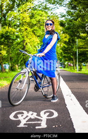 Junge brünette Mädchen im blauen Kleid und Sonnenbrille sitzt auf einem Fahrrad auf einem Fahrrad auf asfalt Straße. Stockfoto