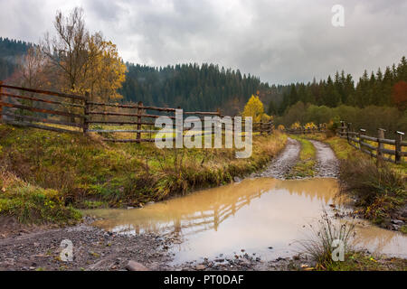 Riesige Pfütze auf dem Land Straße. hölzernen Zaun entlang dem Weg. tiefen Herbst in bergige Landschaft Stockfoto