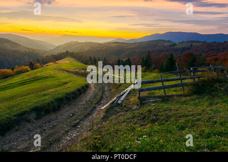 Sonnenuntergang im Herbst Berge. wicklung Land straße durch grasartige ländliche Hügel. hölzernen Zaun entlang dem Weg. Bäume im Herbst Laub. entfernten Ridge in eveni Stockfoto