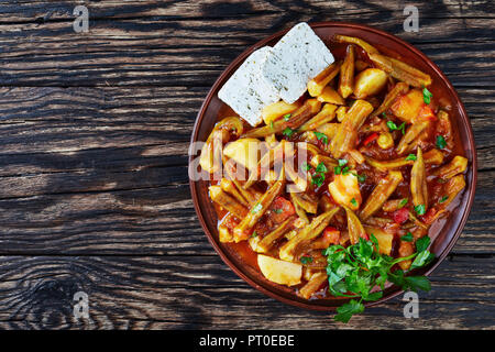Okra Mpamies Giaxni mit Tomaten und Kartoffeln, mit Feta Käse auf einem Steingut Teller auf einem dunklen Holztisch serviert, griechisch Rezept, vi Stockfoto