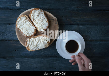 Gesundes Frühstück. Sandwiches mit Sahne, Käse und Kaffee. Ansicht von oben. Schwarzer Holztisch. Tasse Kaffee in der Hand. Stockfoto