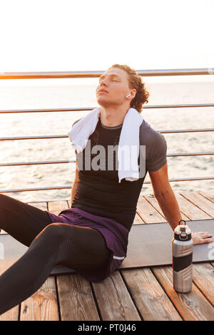 Porträt einer ruhigen Sportler sitzen auf einem Fitness Matte am Strand, Musik hören mit Kopfhörern Stockfoto