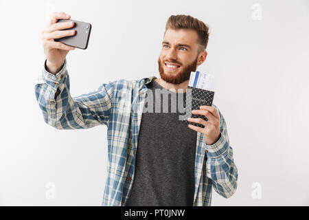 Porträt einer fröhlicher junger bärtiger Mann stehend über Weiß isoliert, mit Reisepass mit fliegenden Karten, wobei selfie Stockfoto