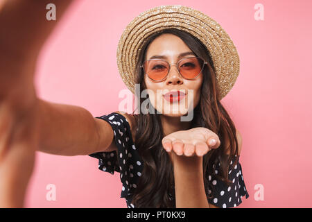 Gerne brünette Frau, Stroh, Hut und Sonnenbrille, selfie während sendet Luft Küssen an der Kamera über rosa Hintergrund Stockfoto