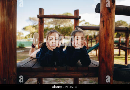 Porträt von zwei kleinen Mädchen glücklich zusammen liegen auf hölzernen Strukturen im Park. Zwillingsschwestern Spaß am Spielplatz. Stockfoto