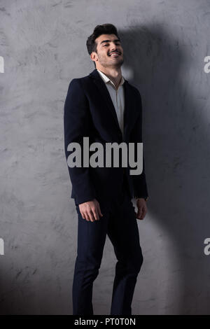 Ein glücklicher hübscher junger Geschäftsmann in einem schwarzen Anzug stand vor einer grauen Wand in einem Studio. Stockfoto