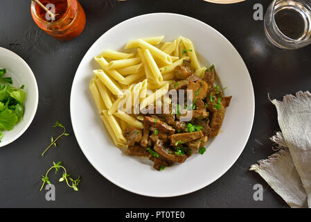 Stroganoff mit Pilzen und Nudeln auf dem Teller über schwarzen Stein. Ansicht von oben, flach Stockfoto