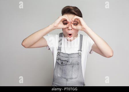 Portrait von überrascht junge brunette Mann im lässigen Stil mit weißen T-Shirt und Jeans Overalls stehen und von der Kamera mit dem Fernglas Geste. Stockfoto