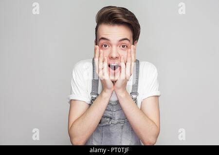 Portrait von erstaunt, schockiert junge brunette Mann in Jeans Overalls stehen und von der Kamera mit großen Augen, offener Mund und sein Gesicht zu berühren. Stockfoto