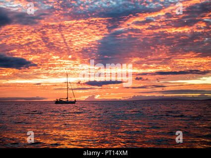 Helles orange Sonnenuntergang Wolken füllen Himmel und auf der Wasseroberfläche mit einem Boot in Silhouette am Horizont spiegeln Stockfoto