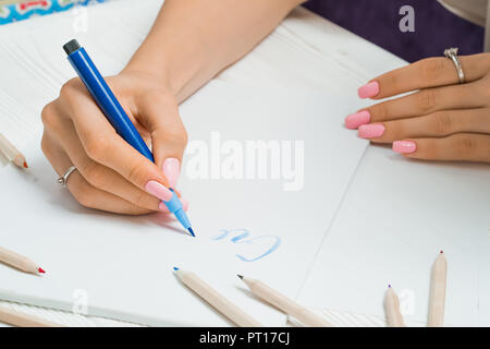 Erstellen. Weibliche Hand schreibt wunderschöne blaue Briefe auf Leinwand in bunten Federn Rahmen. Kalligraphie script. Kunst der Briefe zu schreiben. Frau sitzt auf der alten w Stockfoto