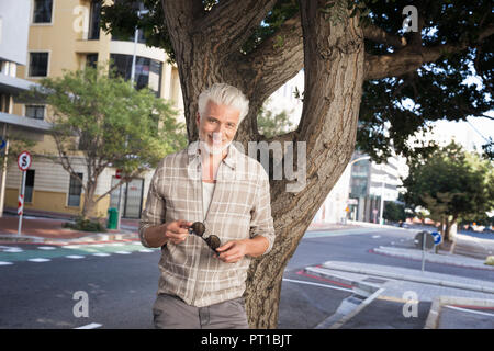 Reifer Mann lehnte sich gegen den Baum, Sonnenbrille Holding Stockfoto