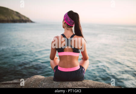 Sportliche Frau sitzt auf Felsen, mit Blick auf das Meer am Abend, Ansicht von hinten Stockfoto