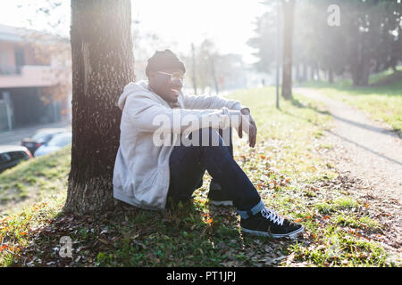 Lachende junge Mann im Park sitzen, lehnen auf Baumstamm Stockfoto