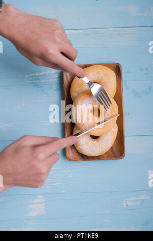 Frühstück Konzept: Nahaufnahme der männlichen Händen mit Gabel und Messer schneiden Donut Form Keks auf Blau Holz- Hintergrund; Ansicht von oben, flach Stockfoto