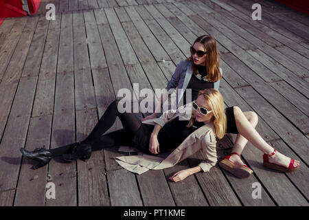 Zwei modische junge Frauen sitzen auf Holzboden mit Sonnenbrille Stockfoto