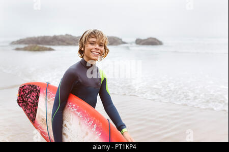 Spanien, Aviles, Portrait von lächelnden jungen Surfer Surfbrett Durchführung am Strand Stockfoto