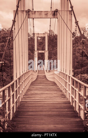 Vintage Suspension Bridge hängen über den Fluss. Alte langen hängenden Holzsteg mit Stahl Schienen über Fluss gegen bewölkter Himmel Hintergrund. Uhr Stockfoto