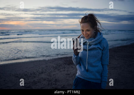 Frau mit Smartphone auf dem Strand bei Sonnenuntergang Stockfoto