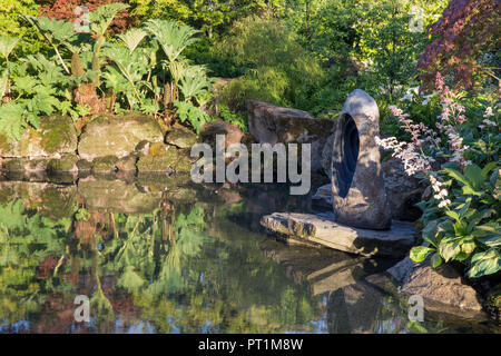 Kleines Teichwasser mit Skulpturen, mit Moos bedeckten Steinen, Felsbrocken, japanischer Garten mit Gunnera manicata Rodgersia aesculifolia UK Stockfoto
