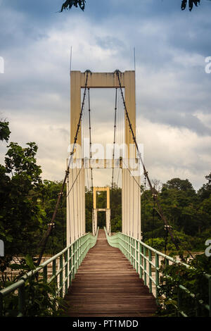 Vintage Suspension Bridge hängen über den Fluss. Alte langen hängenden Holzsteg mit Stahl Schienen über Fluss gegen bewölkter Himmel Hintergrund. Uhr Stockfoto