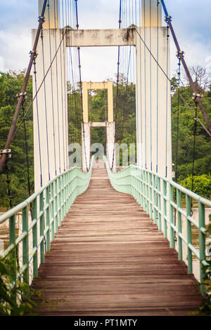 Vintage Suspension Bridge hängen über den Fluss. Alte langen hängenden Holzsteg mit Stahl Schienen über Fluss gegen bewölkter Himmel Hintergrund. Uhr Stockfoto