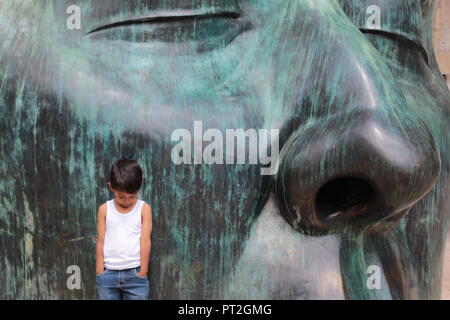 Niño vestido con camiseta Blanca posando Junto eine enorme Escultura de Rostro de Farbe verde en un día Soleado en el centro de Guadalajara Stockfoto