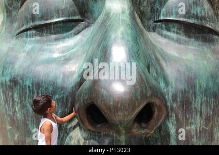 Niño vestido con camiseta Blanca posando Junto eine enorme Escultura de Rostro de Farbe verde en un día Soleado en el centro de Guadalajara Stockfoto