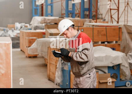 Der Lagerhalter, markiert die Ankunft auf dem Tablett. Mann, der an einem Gabelstapler durch ein Lager in einer Fabrik. Treiber in Uniform und Schutzhelm. Das Konzept der Logistik und Lagerung Stockfoto