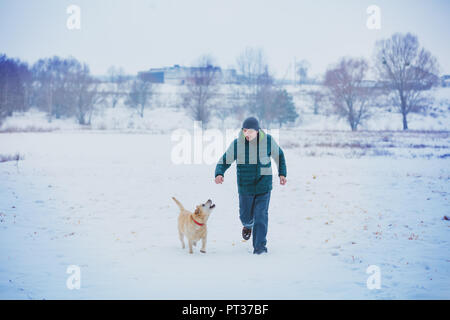 Glückliche Menschen mit einem Labrador Retriever Hund läuft durch ein schneebedecktes Feld im Winter Stockfoto