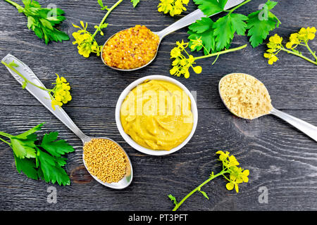 Senf Granulat, Pulver, Samen in Löffel Sauce in eine Schüssel, gelbe Blumen und Petersilie auf dem Hintergrund einer Holzplatte von oben Stockfoto
