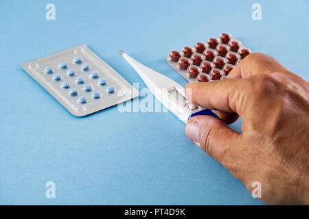 Blisterpackungen mit blau und braun Pillen und Elektronische Digitalthermometer durch einen männlichen Hand auf blauem Hintergrund - Gesundheitswesen und medizinische Konzept Stockfoto