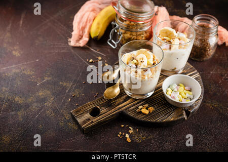 Gesundes Dessert aus Joghurt, eine Banane, Müsli und Nüsse auf einer hölzernen runde Schneidbrett auf einem dunklen Hintergrund Stockfoto