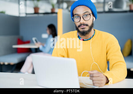 Verdutzt hübscher junger hipster Kerl in Gläsern am Tisch sitzt mit Laptop und hören die Musik im Kopfhörer während Augenbraue und Stockfoto