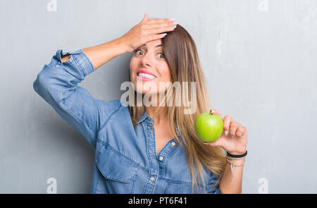 Junge erwachsene Frau über Grau grunge Wand essen frischen grünen Apfel mit Hand betonte am Kopf, mit Scham und Überraschung Gesicht schockiert, wütend und frustriert Stockfoto