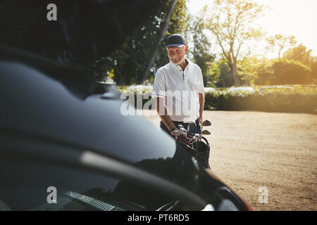 Sportliche älterer Mann lächelnd, während seine Vereine in den Kofferraum seines Autos packen nach dem Spielen Sie eine Runde Golf auf einem sonnigen Nachmittag Stockfoto